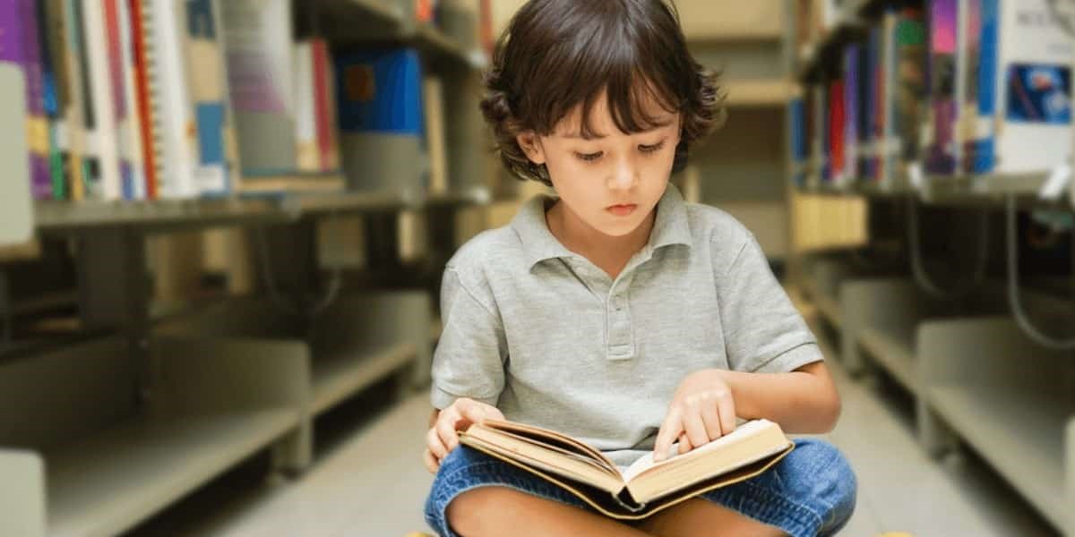 copil care citeste o carte printre rafturile unei biblioteci